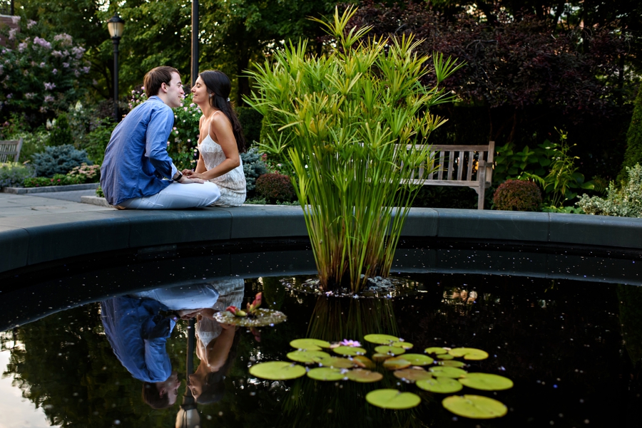 A man and woman sit in battery park next to a small pond at sunset for their engagement session.